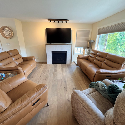 A spacious living room with wide-plank light oak hardwood flooring, tan leather recliners, a modern fireplace with a mounted TV, and neutral-toned decor. The hardwood flooring, installed by Chilliwack Carpet One Floor and Home